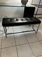 Black faux leather bench with chrome legs on tiled floor near glass table