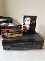 Photo showing Broksonic VHS HQ Video Cassette Recorder with DVDs 24, Six Feet Under, Fat Actress, CSI on top.