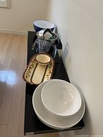 Two white ceramic bowls stacked, rectangular beige ceramic platter with floral design, clear plastic measuring cups stacked inside stainless steel mixing bowls on a black surface.