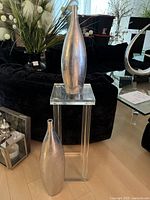 Clear acrylic pedestal table with one silver vase on top and one beside it, displayed on light wooden floor with black furniture in background.
