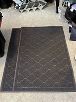 Two dark grey outdoor mats with geometric pattern stacked on carpeted floor