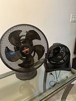 Front view of two black electric fans: larger Comfort Zone fan with five blades and a smaller fan beside it on a table.
