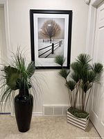 Tall black ceramic vase with artificial palm leaves and grass inside, placed in a corner near a framed artwork.