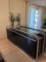 Front and side view of the console table showing the glass top, black cabinetry with three doors, and chrome tubular frame.