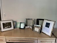 Photo of various picture frames arrayed on a wooden surface showing different sizes including 8x10, 5x7, smaller square and rectangular frames in black, white, silver, chrome, blue, and green colors.