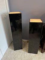 Two black rectangular light pedestals with illuminated amber tops placed against a wall on carpeted floor.