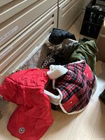 Photo showing several dog jackets and coats, including a red waterproof coat, red plaid fleece jacket, green parka with faux fur, and a black coat, arranged on a floor.
