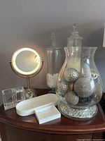 Vanity set arranged on wooden surface featuring lighted mirror, glass apothecary jars, white trays, acrylic organizer, and decorative glass vase with silver spheres.