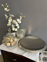 Overall view of the artificial white orchid in silver pot, glass decorative jar filled with natural decor, and large hammered silver tray on a table against a gray wall.