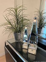 Set of two ribbed clear glass pots each holding an artificial plant with long green leaves, positioned behind two mirror pyramid décor pieces on a glass table.