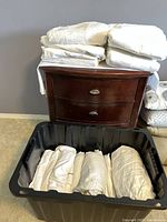 Multiple folded white and cream colored bedsheets stacked on a wooden dresser and some folded inside a black bin.