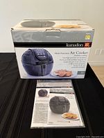 Front view of boxed Kuradori multi function air cooker and recipe booklet in plastic sleeve on table.