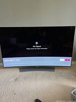 Front view of TV powered on showing 'No Signal' prompt on curved screen with remote on floor.