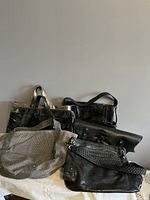 Group of seven handbags stacked together showing variety of styles, colors, and materials including black, grey, and metallic silver with leather and faux leather finishes.