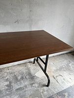 Photo showing the corner of the rectangular brown wooden tabletop and part of the black metal folding leg, table placed on a rough flooring surface.