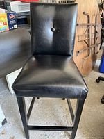 Front view of black faux leather bar stool showing seat, backrest with button tuft, and wooden legs.