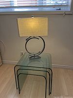 Front view of the smallest clear glass nesting table with the chrome lamp and rectangular shade on top, showing the glass's transparency and rounded corners.