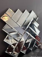 Front angle showing the diamond-shaped, layered arrangement of rectangular mirror panels reflecting a room with furniture and ceiling light fixture.