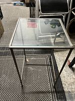 Square side table with clear glass top and silver metal frame, photographed at standing height on a textured floor.