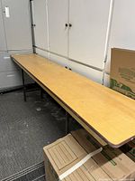 Long rectangular natural wood banquet folding table positioned indoors next to cabinets and cardboard boxes, showing the full length of the table and top surface.