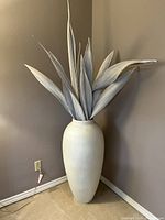 Full view of the tall off-white plaster vase with neutral toned dried palm leaves from the front corner in a beige room corner.