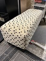 Side angle view of rectangular beige fabric ottoman with black and brown polka dots and dark wooden legs.