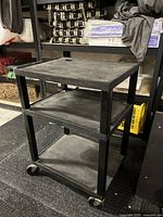 Side and angled view of a black plastic utility cart with three shelves and four wheels, showing surface scratches and dust.