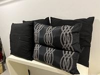 Five pillows grouped together showing two rectangular embroidered pillows placed on top of three plain square black pillows