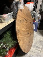 Round wooden table top with natural wood grain and knots, some scratches visible, captured leaning upright against a shelf in a garage.