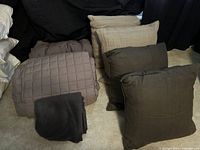 Photo showing four large throw pillows (two dark grey, two beige), two folded king size comforters in dark grey, and one dark grey throw blanket, all placed on carpeted floor.