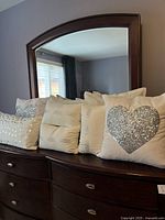 Image showing multiple white cotton pillows with silver sequined hearts, polka dots, and stripe patterns arranged on a dark wooden dresser beneath a mirror in a room with grey walls.