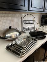 Photo showing stainless steel wok pan with lid, set of 5 stainless steel nested trays, metal two-tier wire stand, cast iron pan with lid with black handle