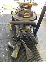 Stanley 8 gallon wet/dry vacuum with attached hose, nozzle, and crevice tool on ground demonstrating included accessories.
