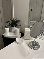 Full view of the set showing the soap dispenser, toothbrush holder, artificial plant, soap dish, clear container, and magnifying mirror on a white surface near a mirror and wall.