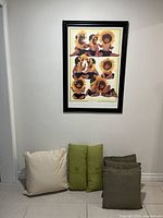 Framed print on wall with three cushions placed on floor: beige cushion, two green cushions, and brown cushions stacked.