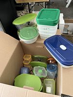 Overview of multiple Tupperware plastic food storage containers inside and on top of a cardboard box, showing variety of shapes and lid colors.
