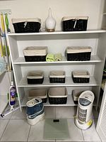 Full view of white shelving unit holding eight black wicker baskets with removable cream fabric liners in various sizes. On floor beneath shelf: Vicks humidifier, Honeywell air purifier, Bios scale, and Swiffer mop.