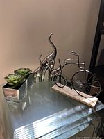 Photo showing the medium vintage-style decorative bicycle model on a wooden base beside two chrome elephant sculptures and a small green plant in a silver rectangular planter on a glass table.