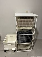 Front view of the wire metal storage rack containing two cream wire baskets and one black fabric basket, alongside the cream rolling bin on wheels
