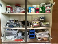 Full view of cabinet with open doors showing three shelves of assorted hardware including screws, bolts, plastic boxes, hose clamps, and electrical conduit boxes.
