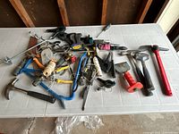 Photo of assorted hand tools spread on a white table showing various hammers, clamps, pliers, garden hoe, and wire rolls.