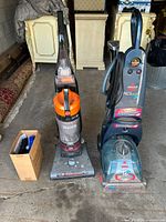 View of Bissell ProHeat 2X carpet cleaner next to Hoover WindTunnel vacuum cleaner and a box of accessories, front angle.