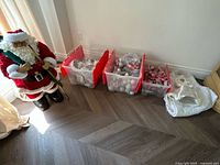 Full lot view showing large Santa figurine, three red-lidded plastic bins filled with Christmas ornaments, and a white Christmas tree skirt.
