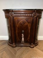 Front view of curved wooden display cabinet with carved detailing, glass door with oval inlay, and decorative handle with tassel.