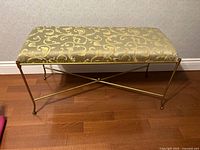Front view of decorative bench showing gold and beige patterned upholstered seat and metal gold-tone frame with curved legs.