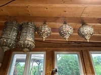 Photo showing all five Moroccan style hanging lanterns suspended from a wooden ceiling beam, including two cylindrical lanterns on the left and three globe-shaped lanterns on the right.