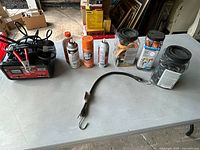 Full view of all garage items on table including battery charger, spray cans, jars with electrical supplies, and steel strap.
