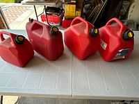 Five red plastic gas cans of varying size on a white table in a garage space, each with black and yellow caps, showing full set and condition.
