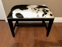 Front view of a black and white hallway bench with patchwork cowhide upholstery and black wooden legs on a wooden floor.