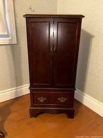 Closed view of the tall wooden jewelry box with front doors showing dark polished wood, golden handles, and a bottom drawer with two ornate gold handles.